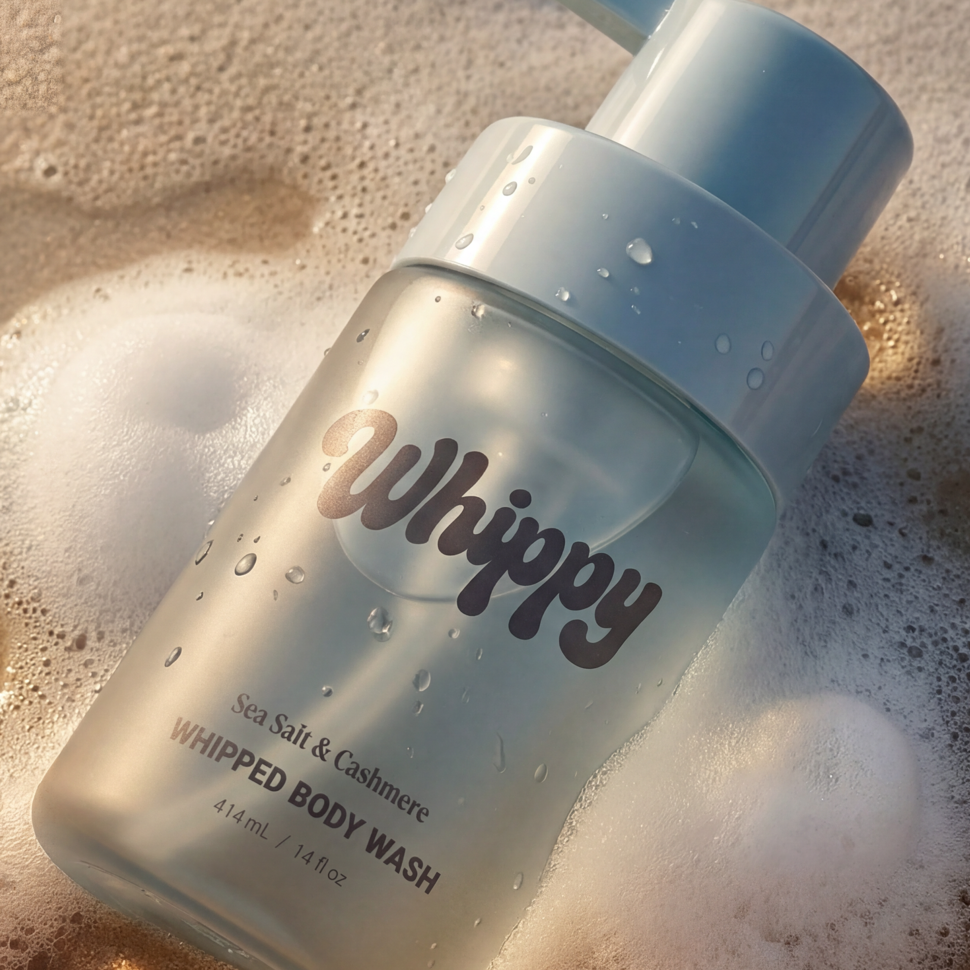 Whippy Sea Salt & Cashmere Whipped Body Wash bottle with water droplets resting on wet sand with foam lather